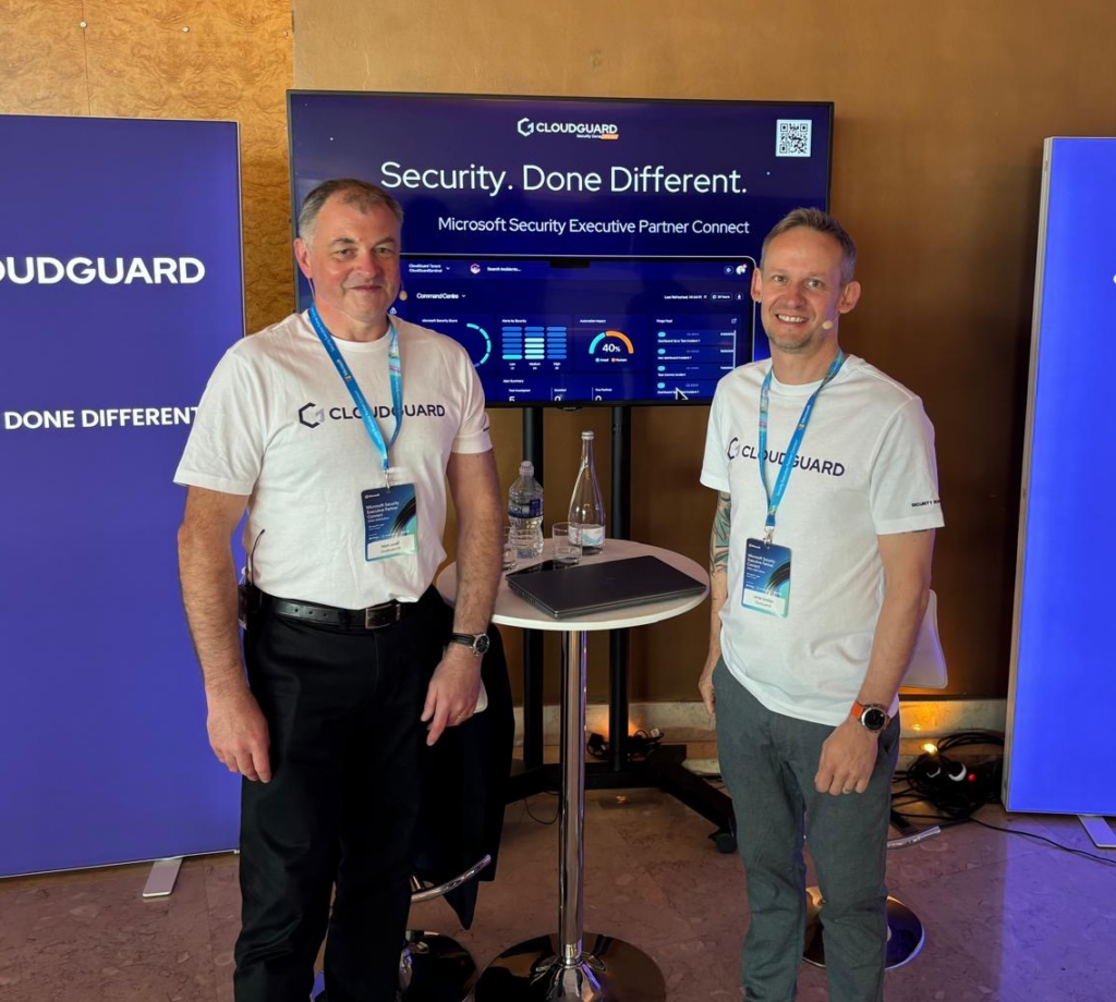 Two men stood in front of banners at security conference wearing white cloudguard t-shirts
