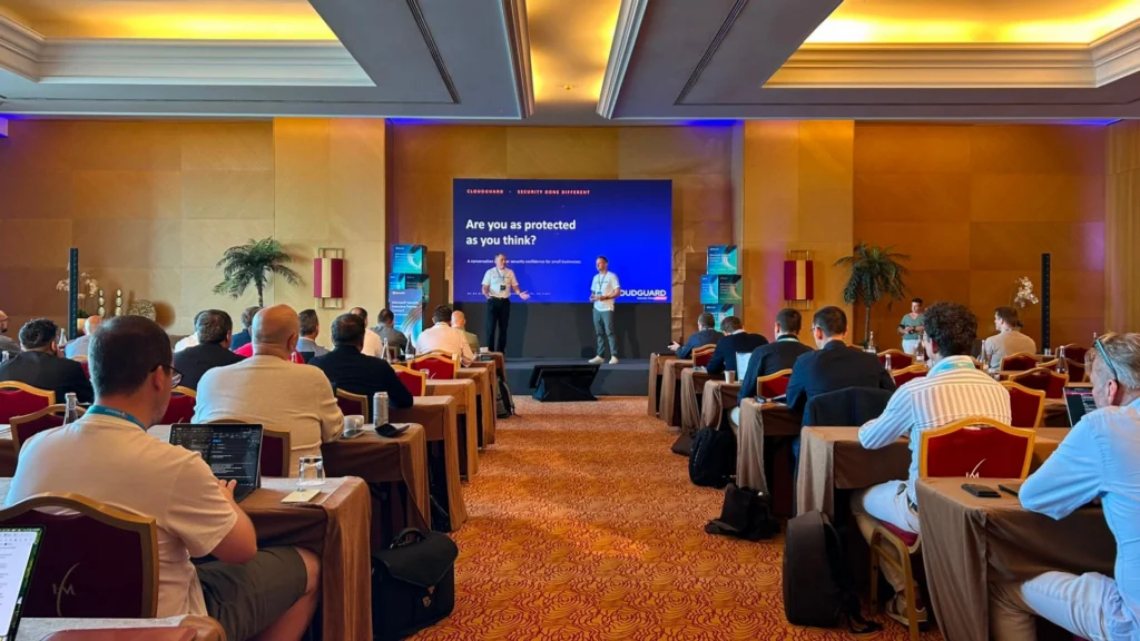 Room filled with people at conference and two people presenting on a screen